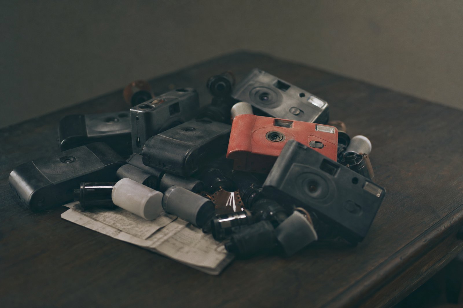 A heap of used disposable film cameras and spent film canisters on a dark table, resting on a folded receipt