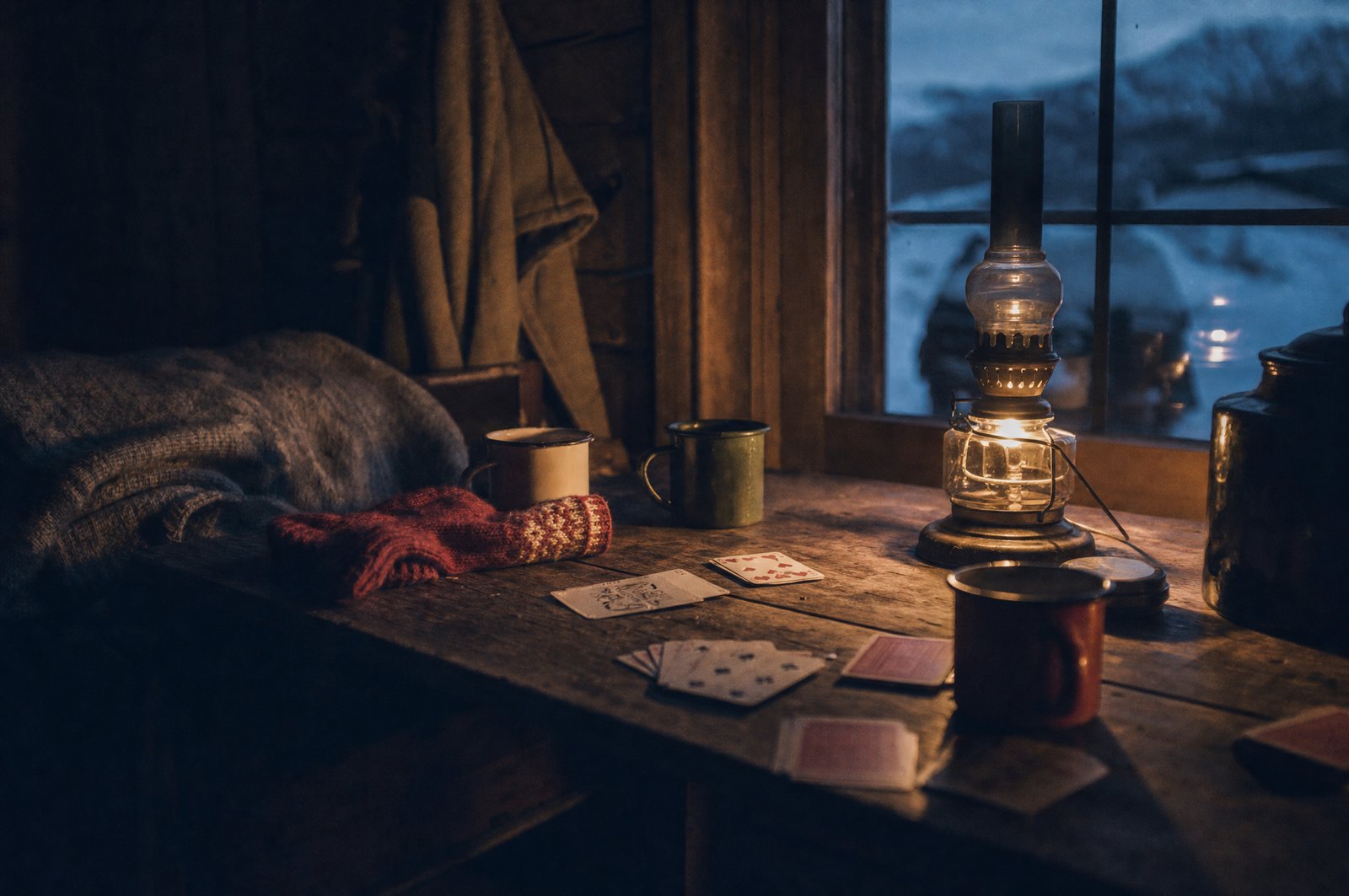 Et slitt trebord ved hyttevinduet med parafinlampe, emaljekopper og kortstokk, vinterskumring utenfor